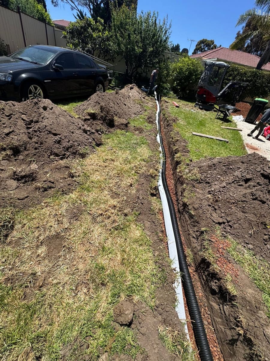 A narrow trench running the length of a back lawn with slotted ag pipe bedded in red scoria and wrapped in white geotextile.
